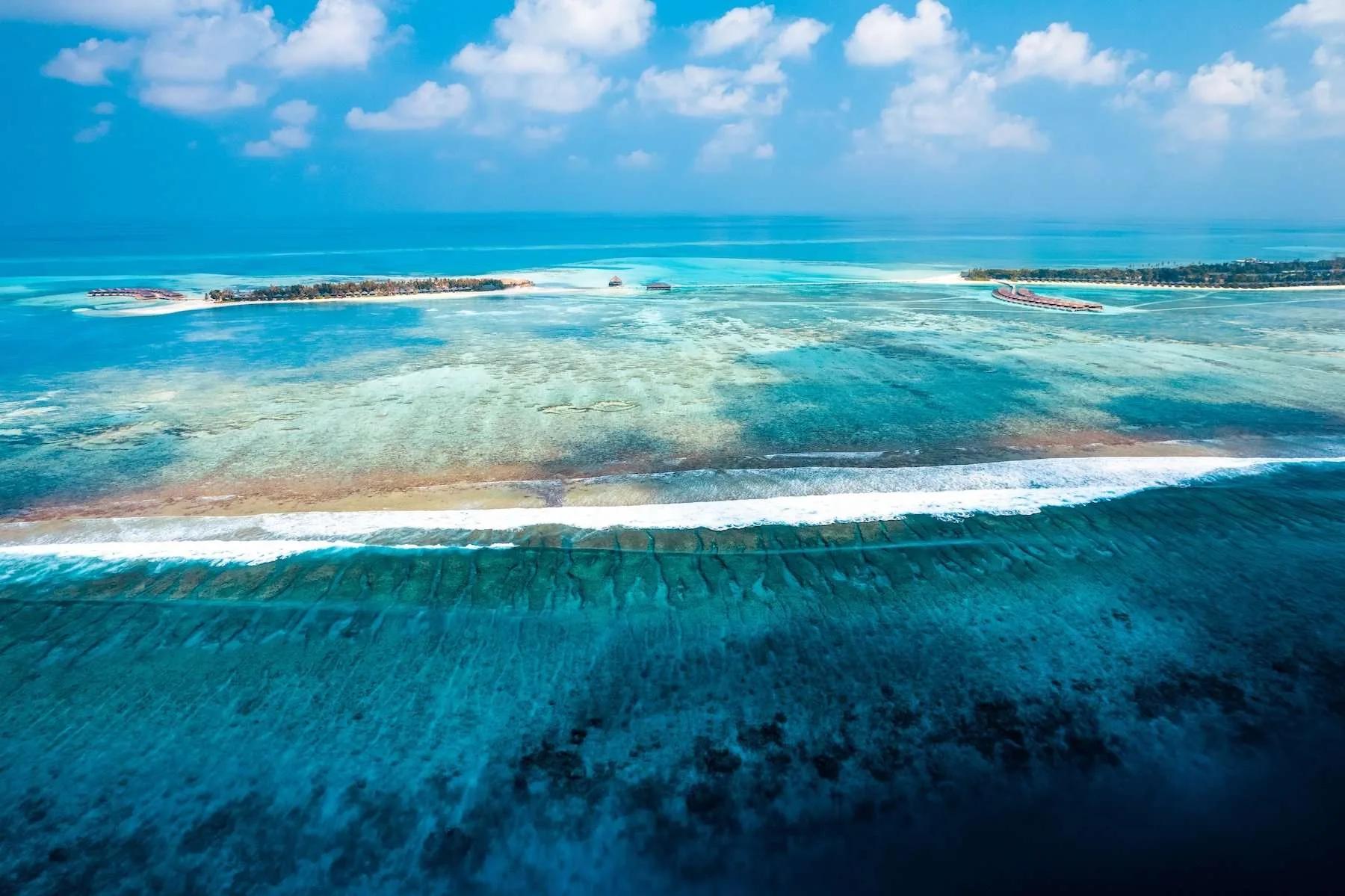 Aerial view of the reef line between Jawakara's two islands, Mabin visible in the distance to the left and Dheru to the right, waves breaking over the barrier reef in the foreground