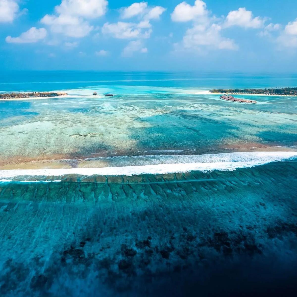 Aerial view of the reef line between Jawakara's two islands, Mabin visible in the distance to the left and Dheru to the right, waves breaking over the barrier reef in the foreground