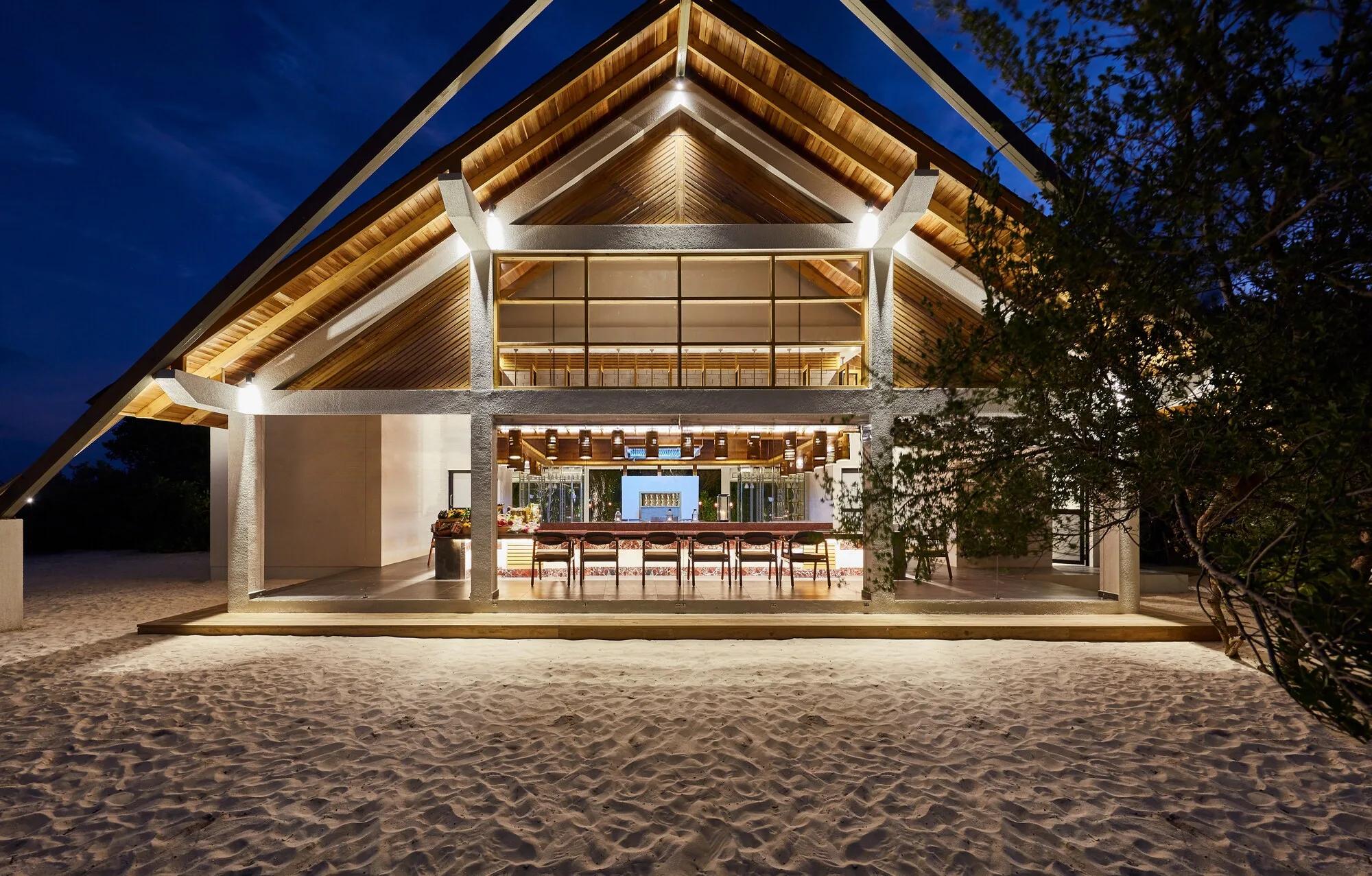 SURF restaurant's timber-framed pavilion lit up at dusk, its open kitchen and bar seating visible through floor-to-ceiling glass beneath a peaked thatched-style roof