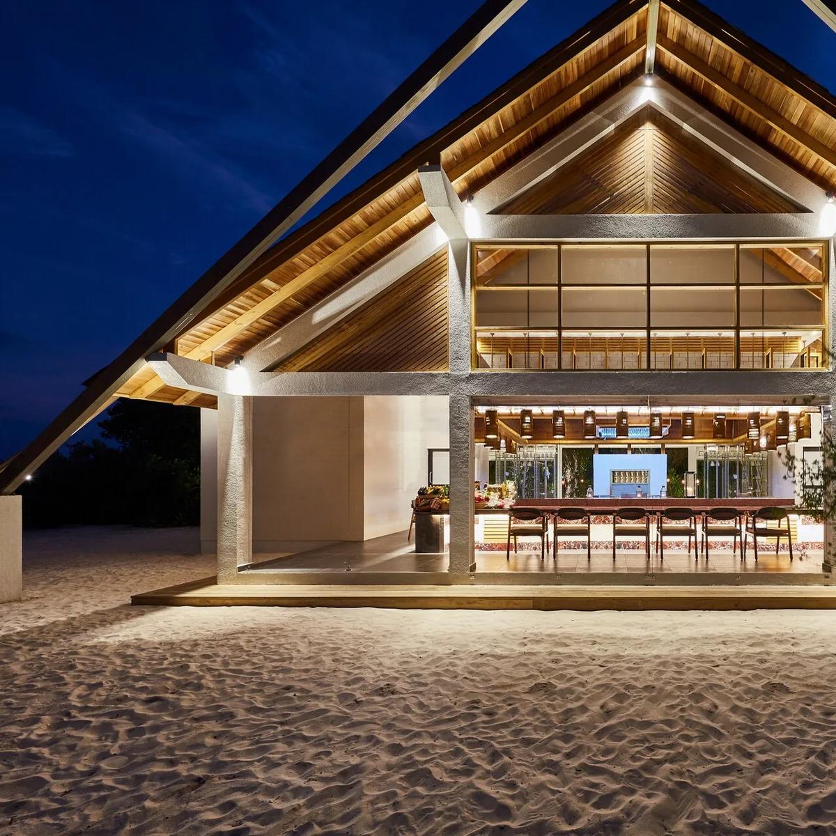SURF restaurant's timber-framed pavilion lit up at dusk, its open kitchen and bar seating visible through floor-to-ceiling glass beneath a peaked thatched-style roof