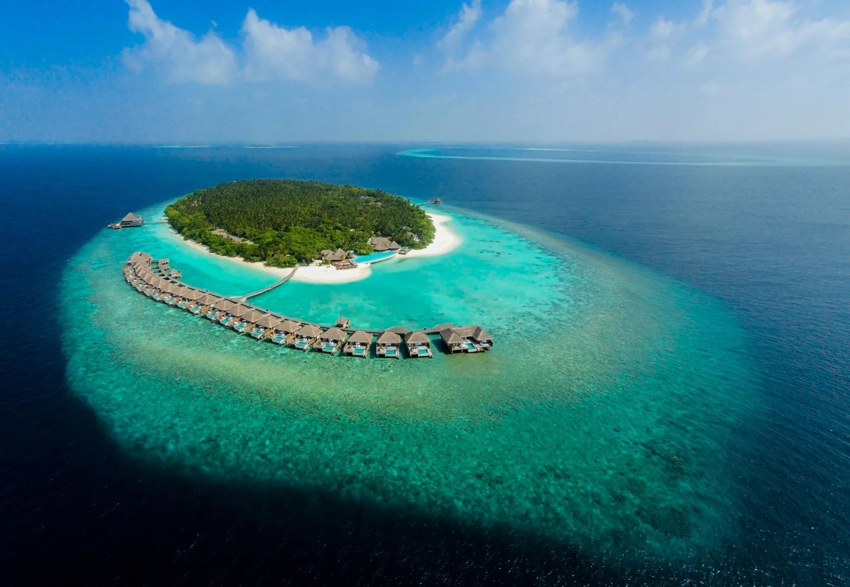 A wider aerial of Dusit Thani Maldives' island and reef, the overwater villa row visible along the western edge