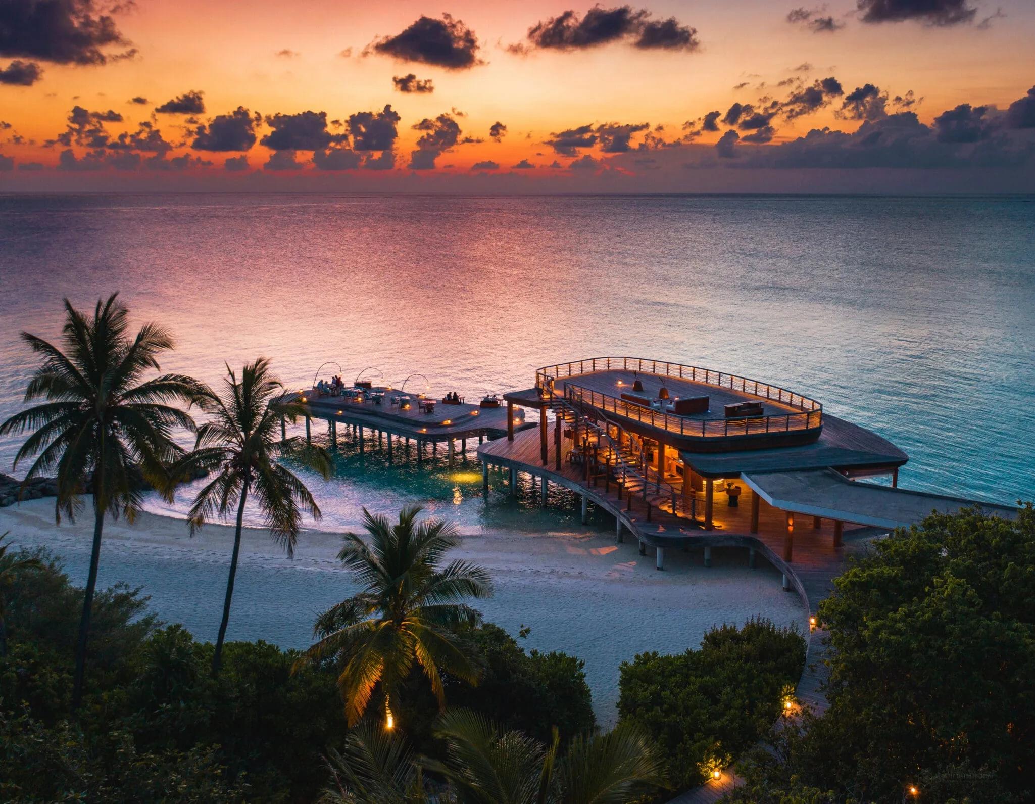 Aerial view of Kata's curved overwater dining pavilion and rooftop bar deck, lit at dusk with the lagoon around it