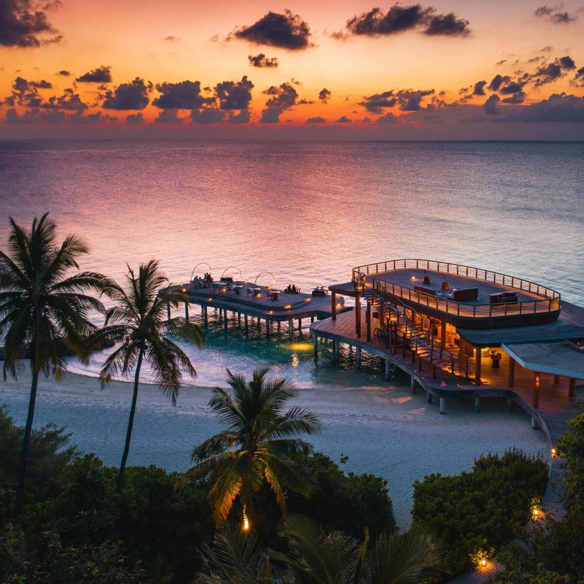 Aerial view of Kata's curved overwater dining pavilion and rooftop bar deck, lit at dusk with the lagoon around it