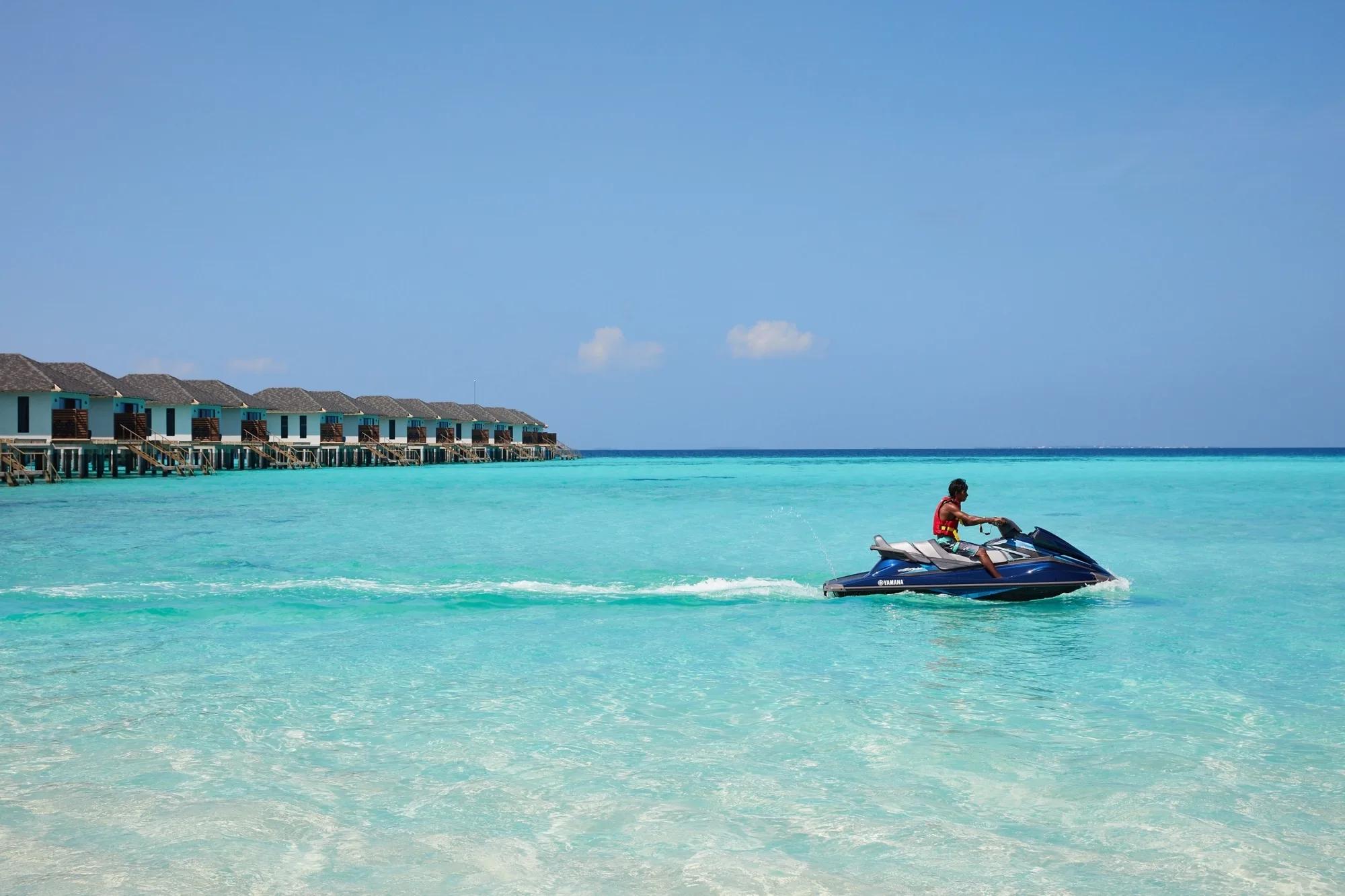 A resort watersports instructor riding a jet ski across the lagoon, a row of overwater villas lined up on the horizon behind him