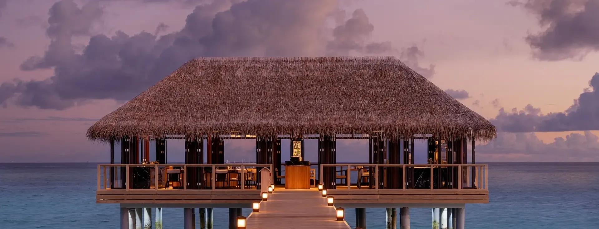 Sora, the overwater restaurant, silhouetted at dusk with its thatched roof over the lagoon