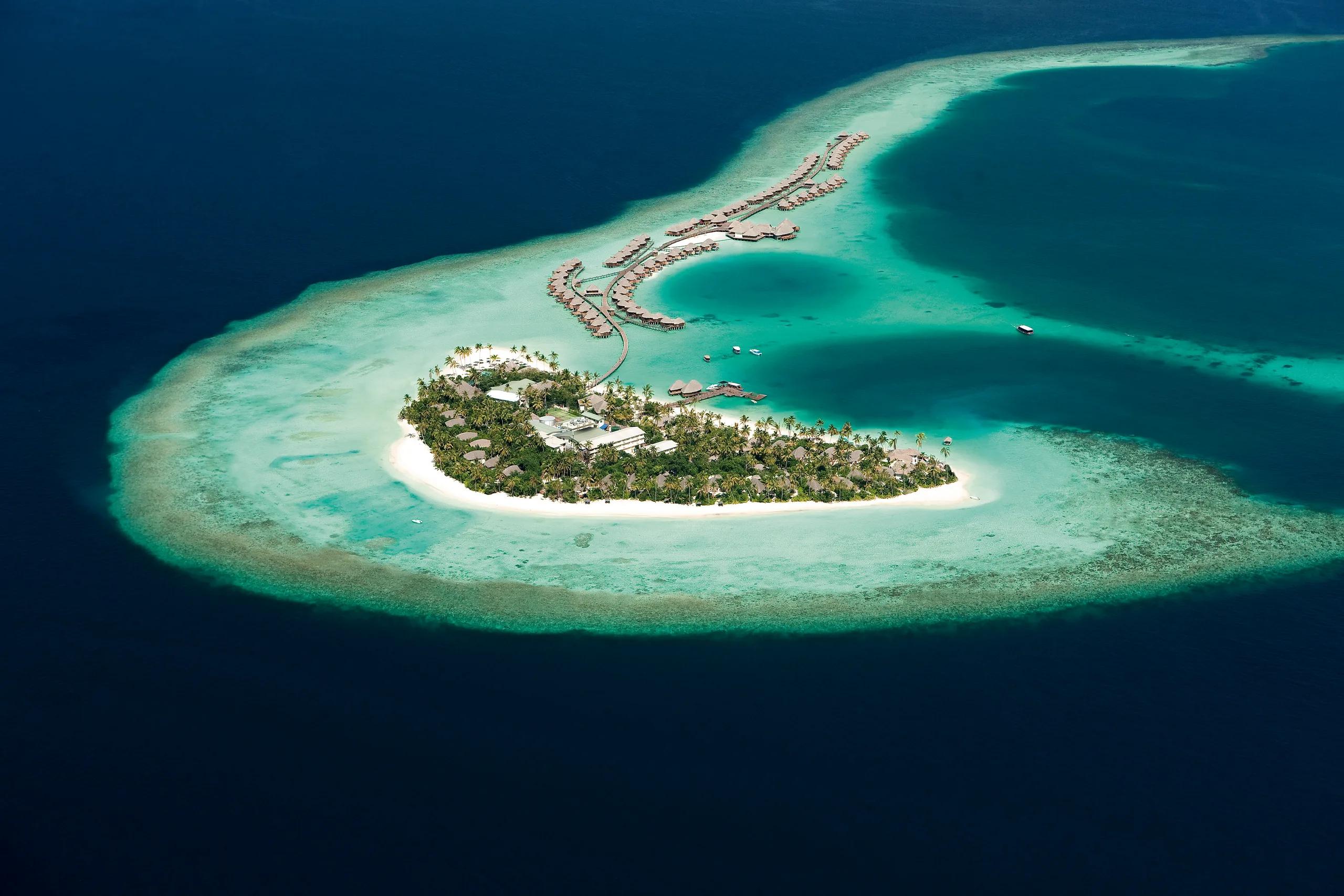 Aerial view of Constance Halaveli's Dhoni-shaped island, ringed by reef, with the resort's overwater villas strung along a long jetty curving into North Ari Atoll's lagoon