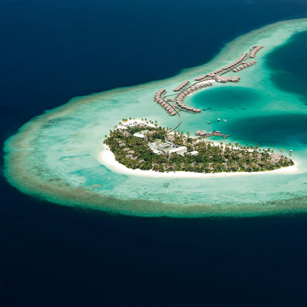 Aerial view of Constance Halaveli's Dhoni-shaped island, ringed by reef, with the resort's overwater villas strung along a long jetty curving into North Ari Atoll's lagoon