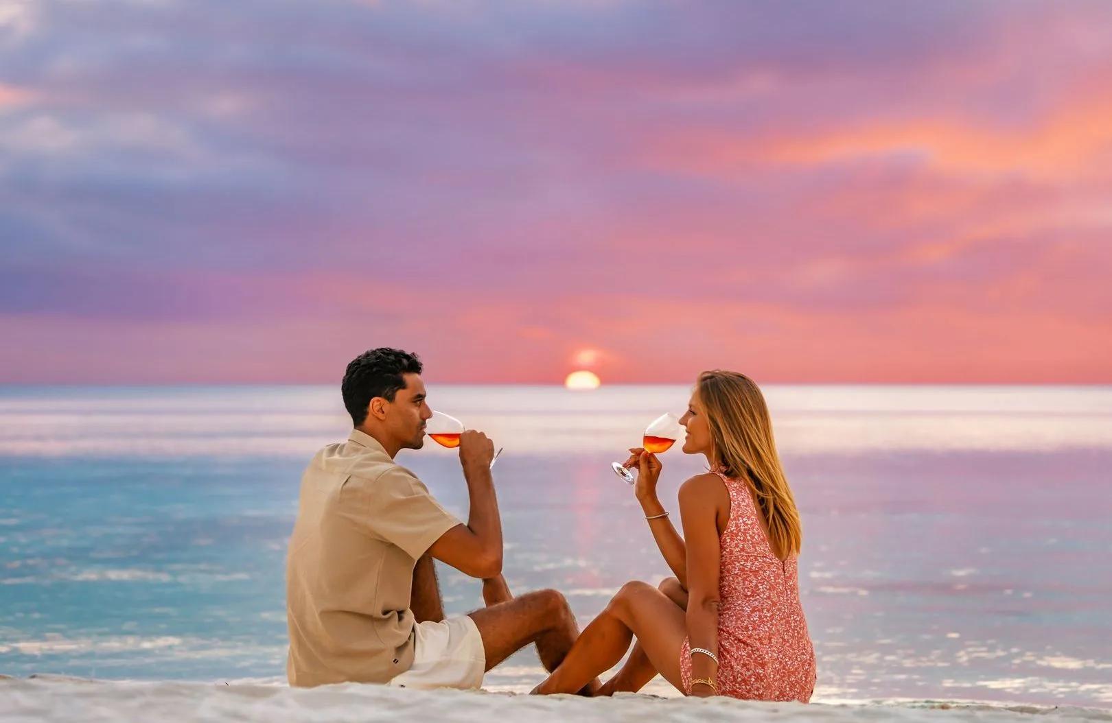A couple seated on the beach at sunset, each holding a glass of rosé, the sun low over the water