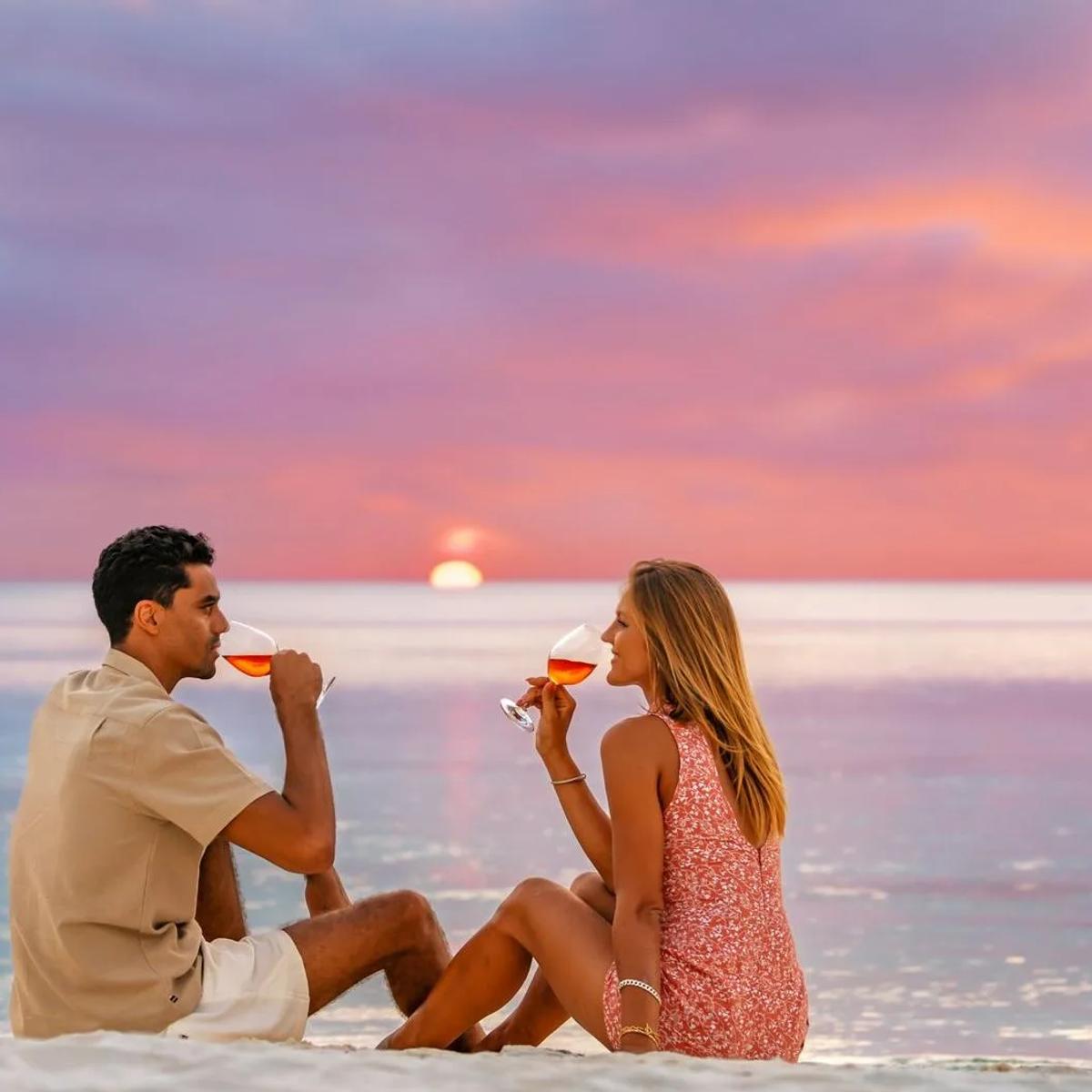 A couple seated on the beach at sunset, each holding a glass of rosé, the sun low over the water