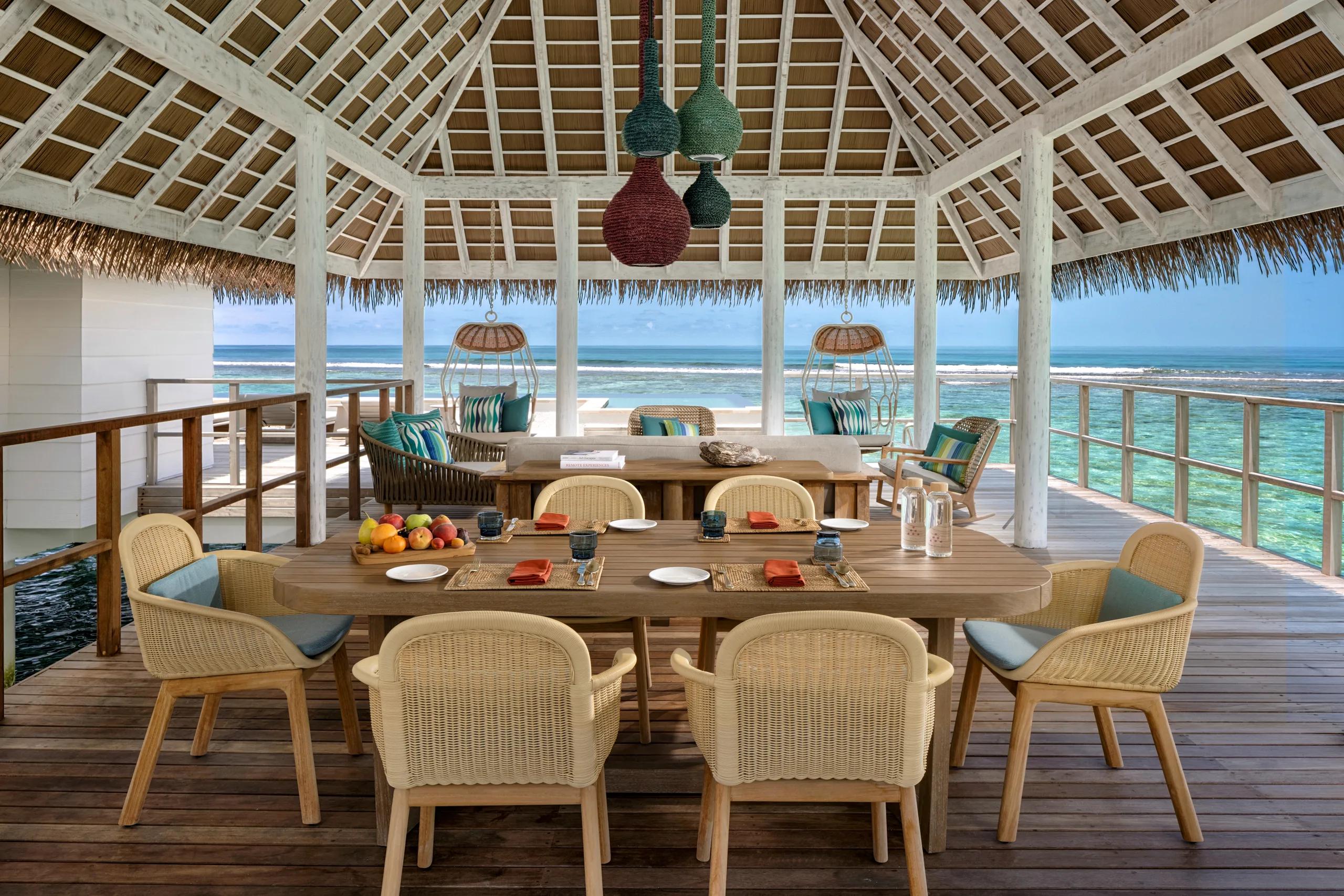 The overwater dining terrace of a Two Bedroom Water Villa with Pool, with wicker furniture, hanging chairs, and an open ocean view.