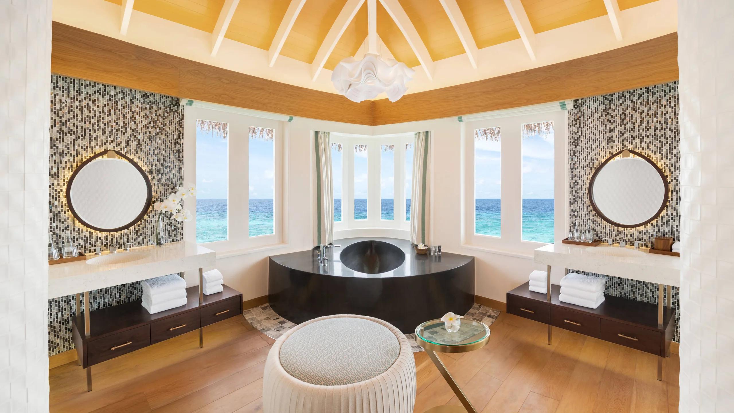 The bathroom of an Overwater Pool Villa, a freestanding tub set beneath a peaked thatch ceiling with lagoon views on three sides