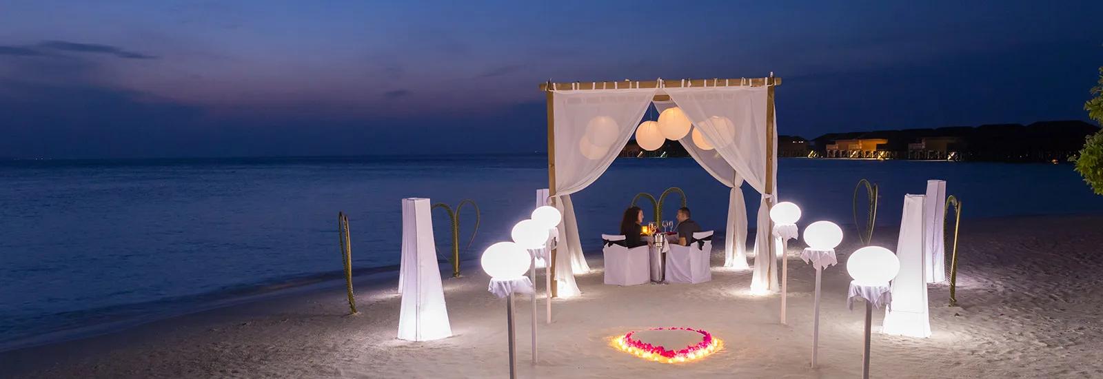 A private dinner table set beneath a draped pavilion on Vilamendhoo's beach at dusk