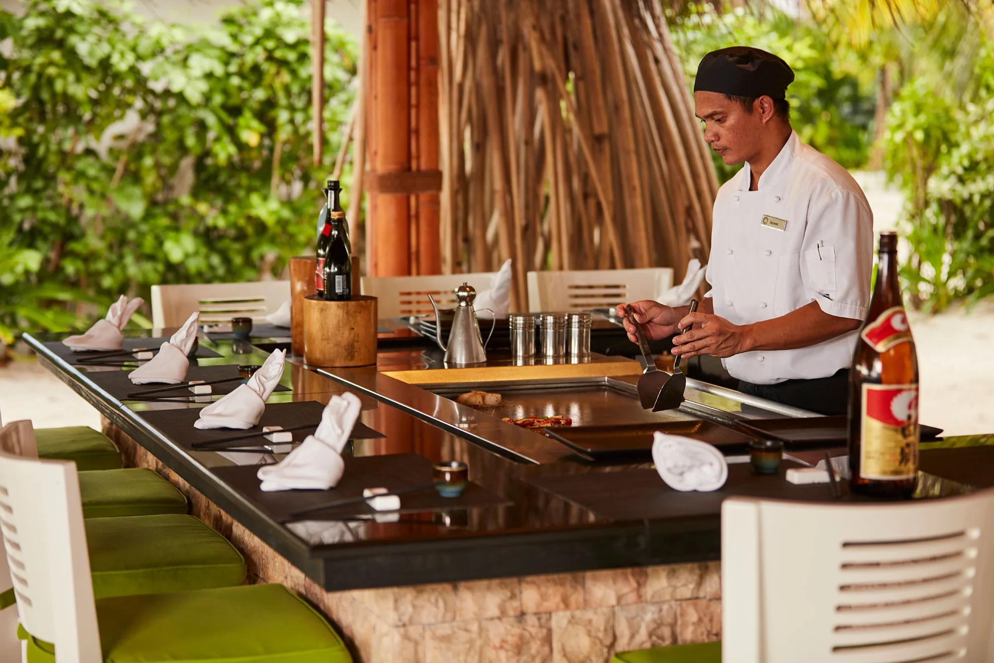A chef in whites cooking table-side at the iron griddle of Teppanyaki, set beneath a thatched canopy