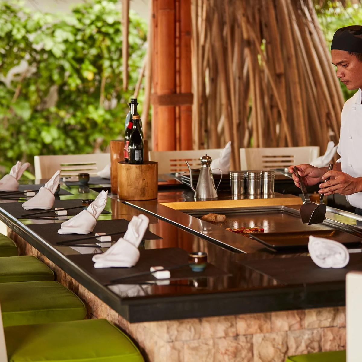 A chef in whites cooking table-side at the iron griddle of Teppanyaki, set beneath a thatched canopy