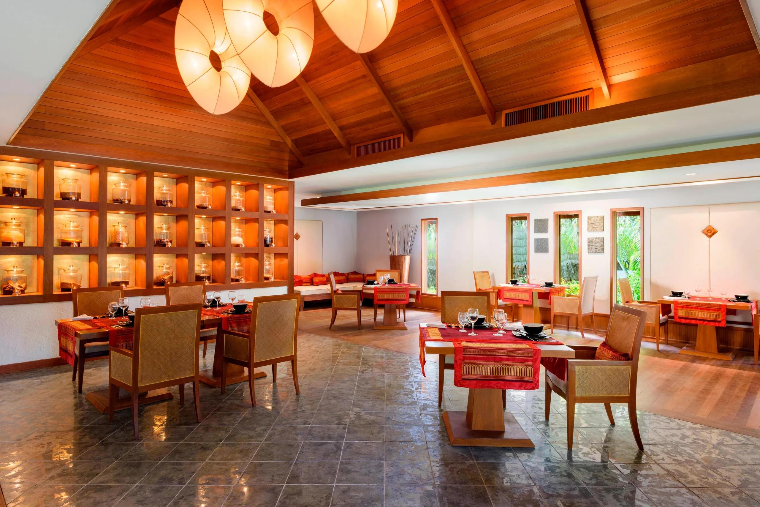 An indoor pavilion dining room with a vaulted timber ceiling, a lit glass-jar display wall, and red table settings
