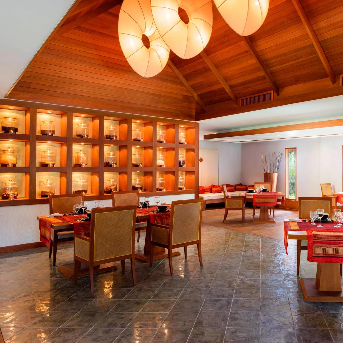 An indoor pavilion dining room with a vaulted timber ceiling, a lit glass-jar display wall, and red table settings