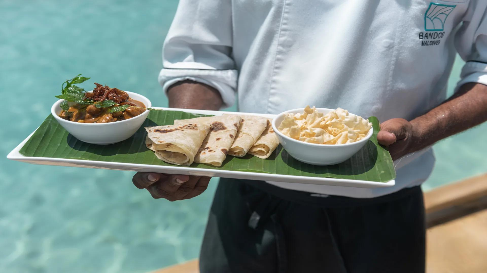 A chef presenting a Maldivian curry with rolled roti and papadum on a banana-leaf tray at Sea Breeze, the resort pool behind him