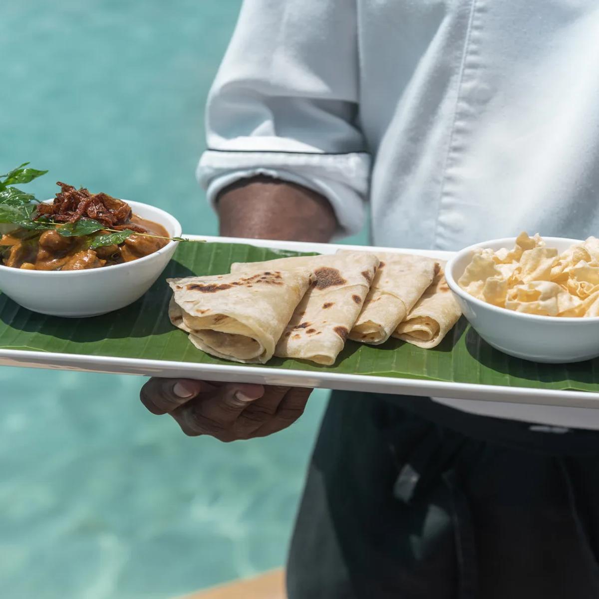 A chef presenting a Maldivian curry with rolled roti and papadum on a banana-leaf tray at Sea Breeze, the resort pool behind him