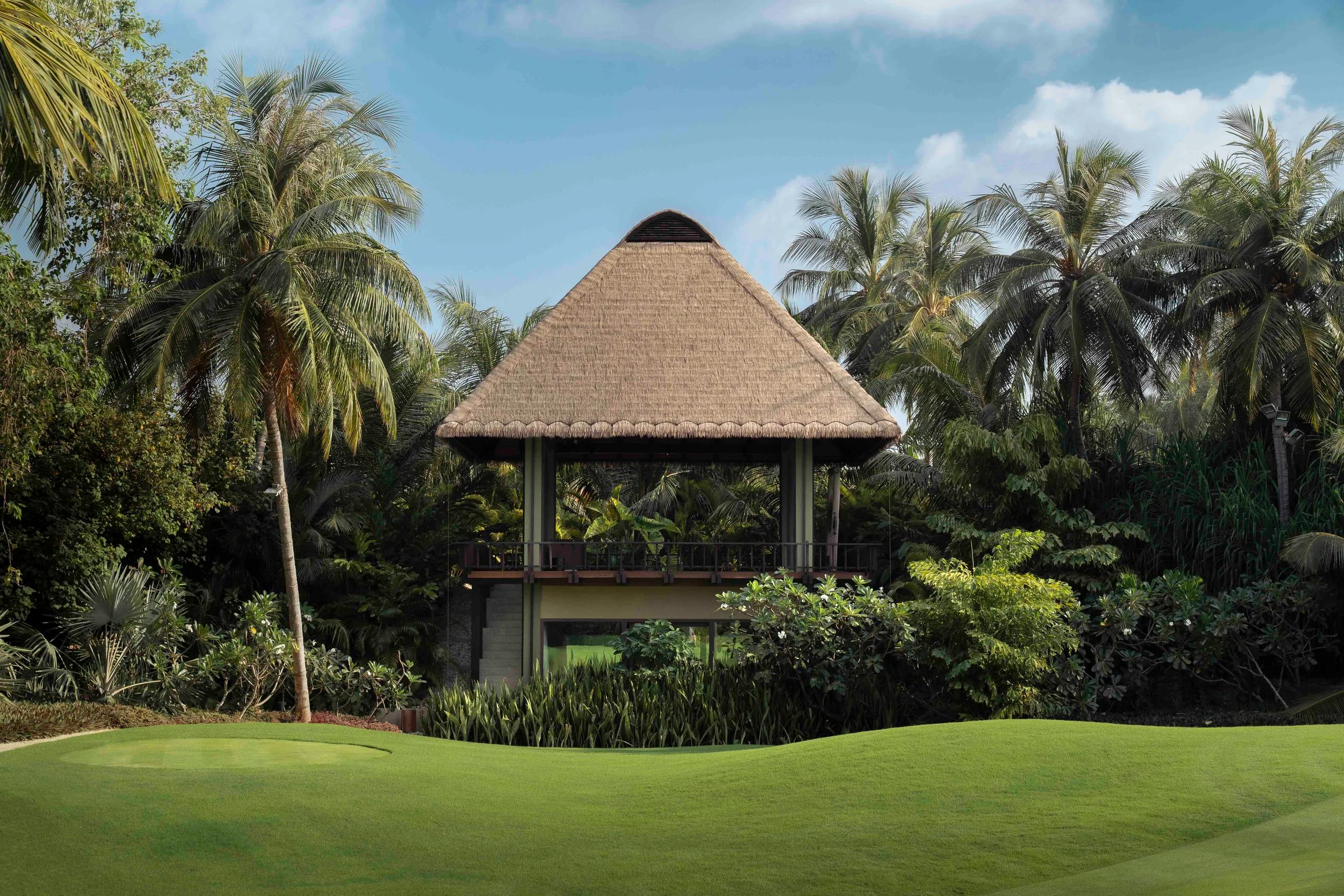 A thatched two-storey clubhouse overlooks a putting green at the Velaa Golf Academy, palm trees framing the building