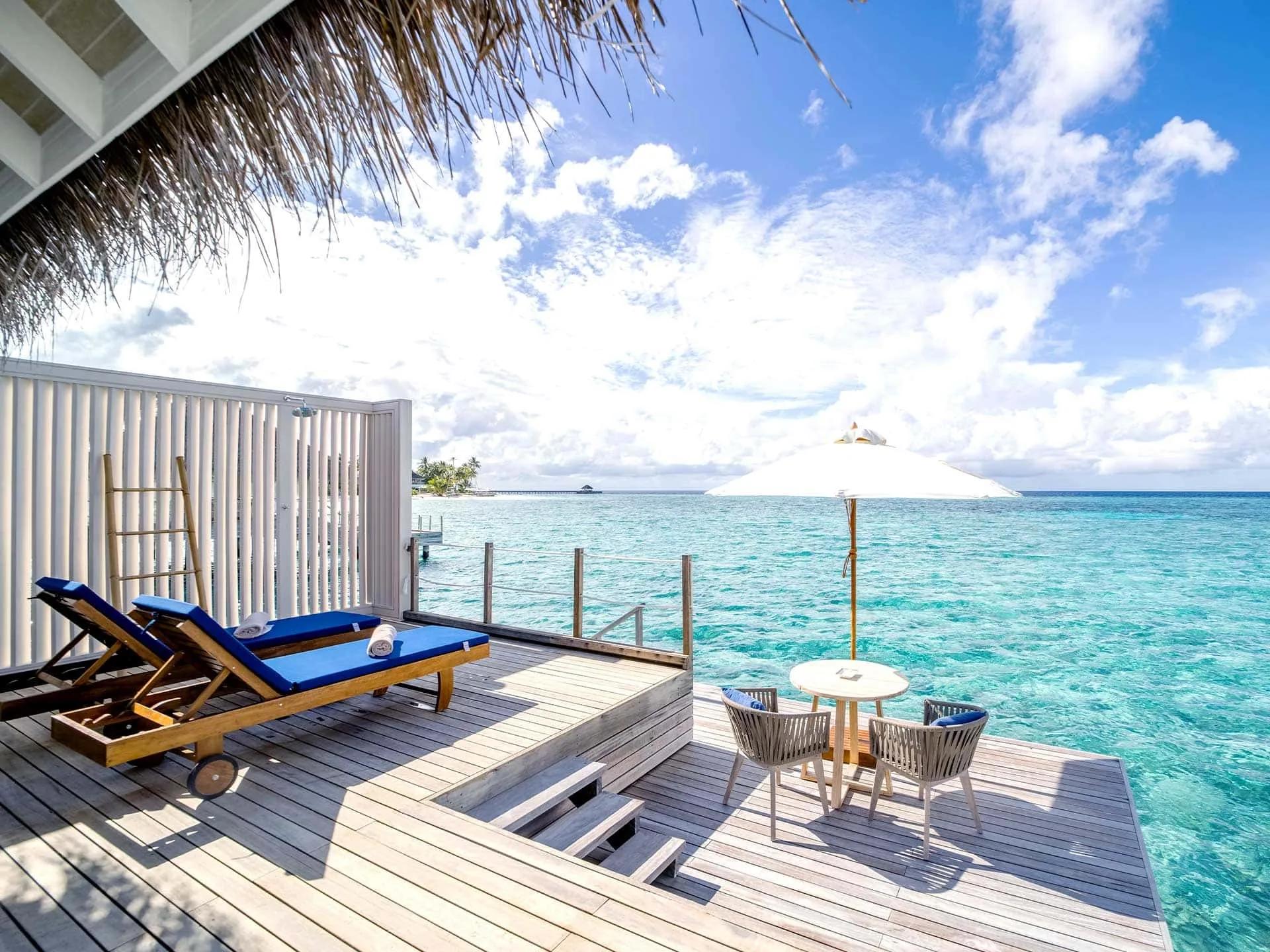 Sun loungers and a table for two on a Water Villa's overwater deck at Baglioni Maldives, with a neighbouring island on the horizon
