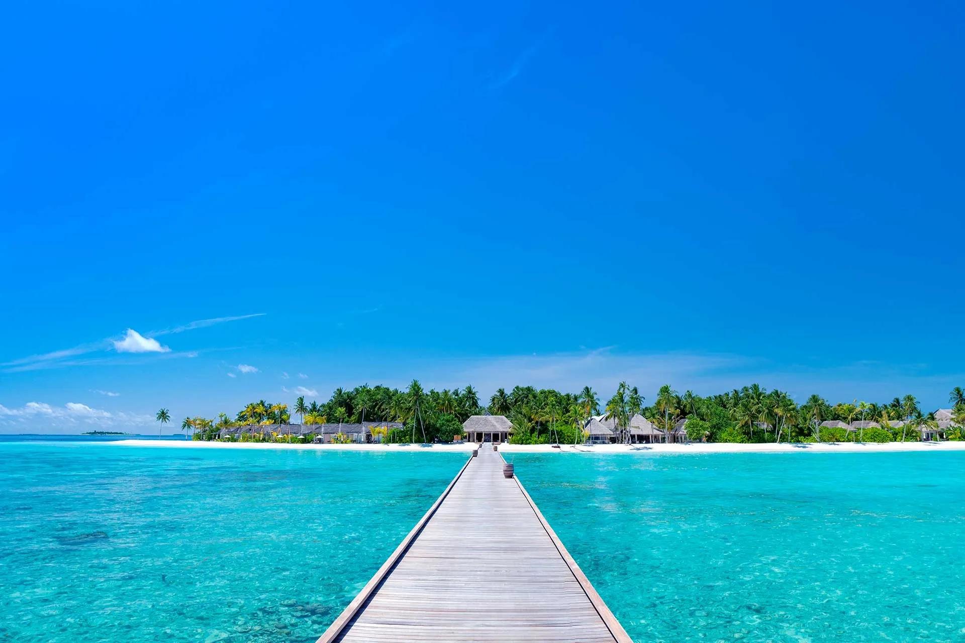 A wooden jetty leading from open lagoon water toward Maagau Island's beach and thatched-roof buildings
