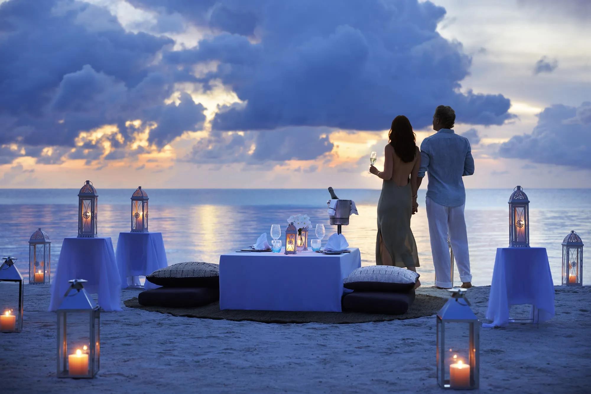 A couple standing at the water's edge beside a private beach dinner table lit by lanterns at dusk