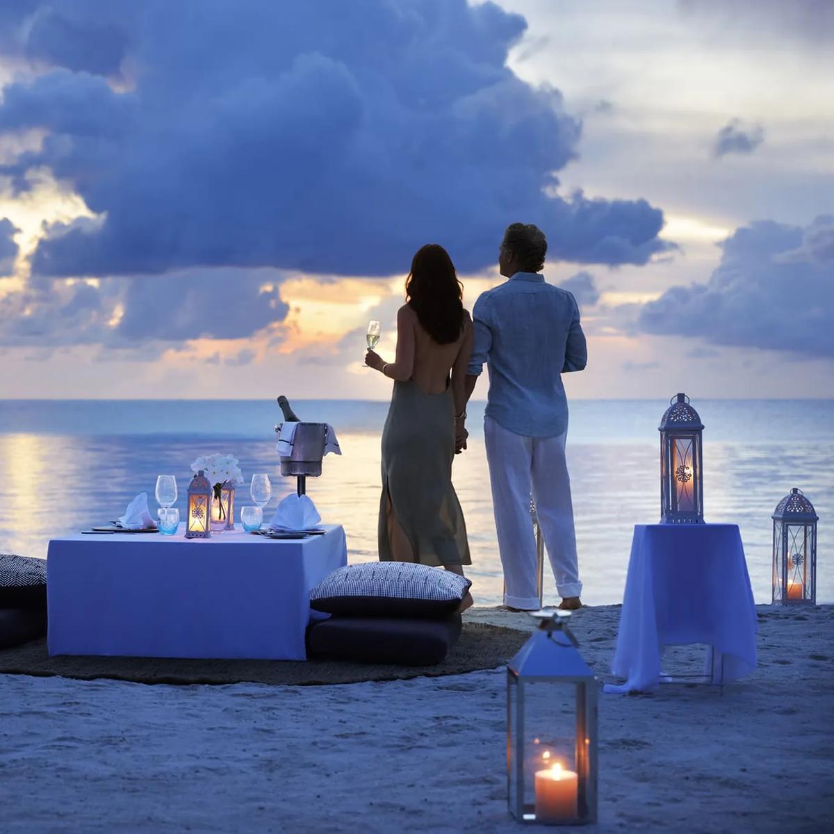 A couple standing at the water's edge beside a private beach dinner table lit by lanterns at dusk