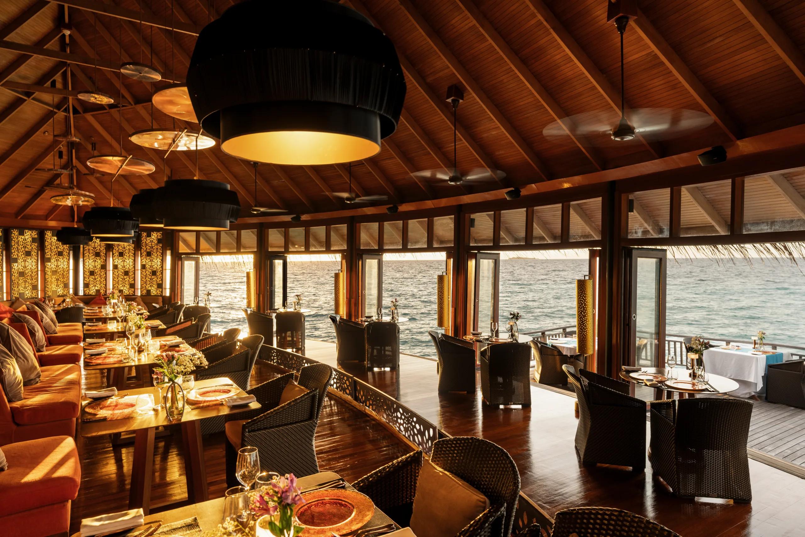 The interior of Jing restaurant at dusk, orange banquette seating and pendant lamps beneath a thatched roof, tables set beside floor-to-ceiling windows onto the ocean