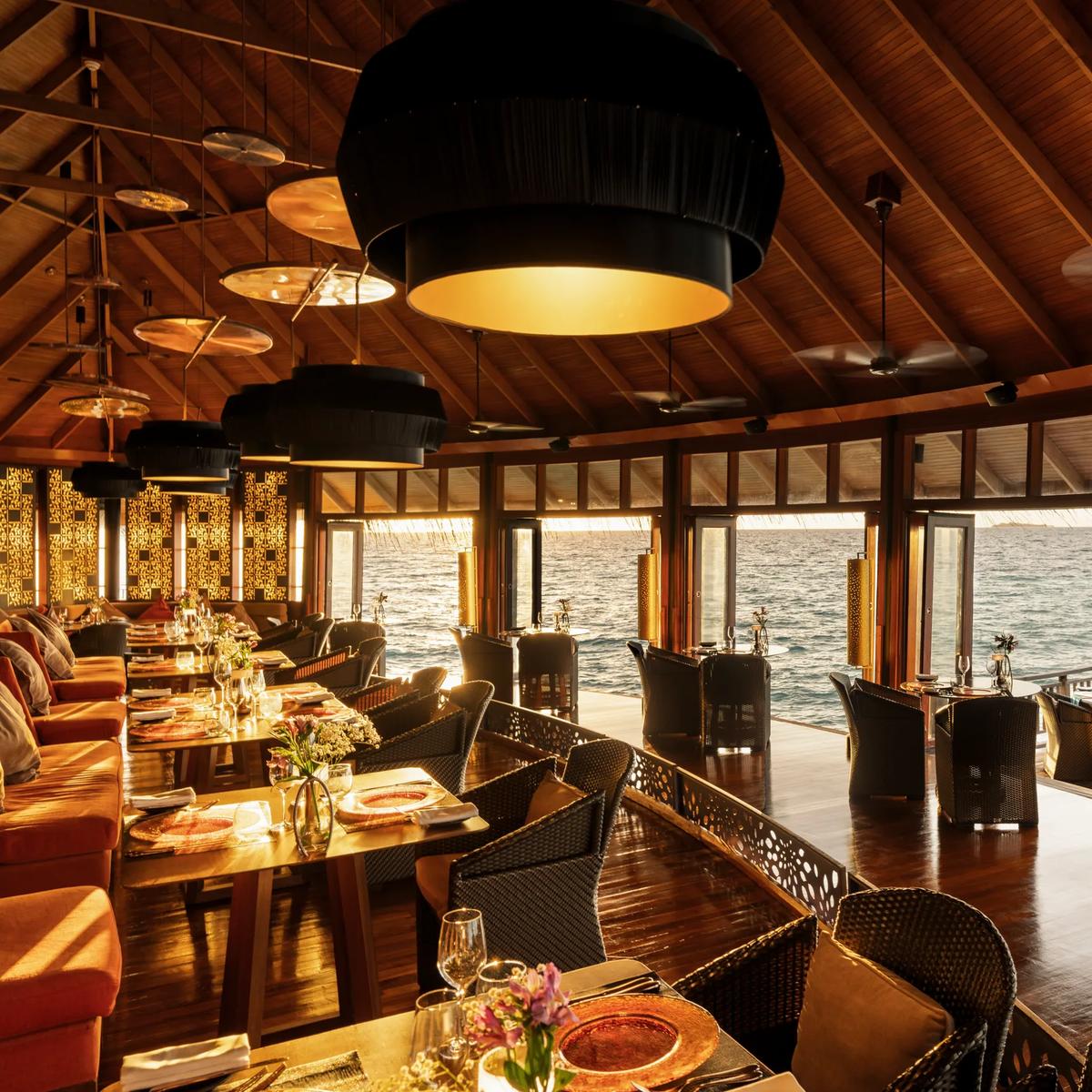 The interior of Jing restaurant at dusk, orange banquette seating and pendant lamps beneath a thatched roof, tables set beside floor-to-ceiling windows onto the ocean
