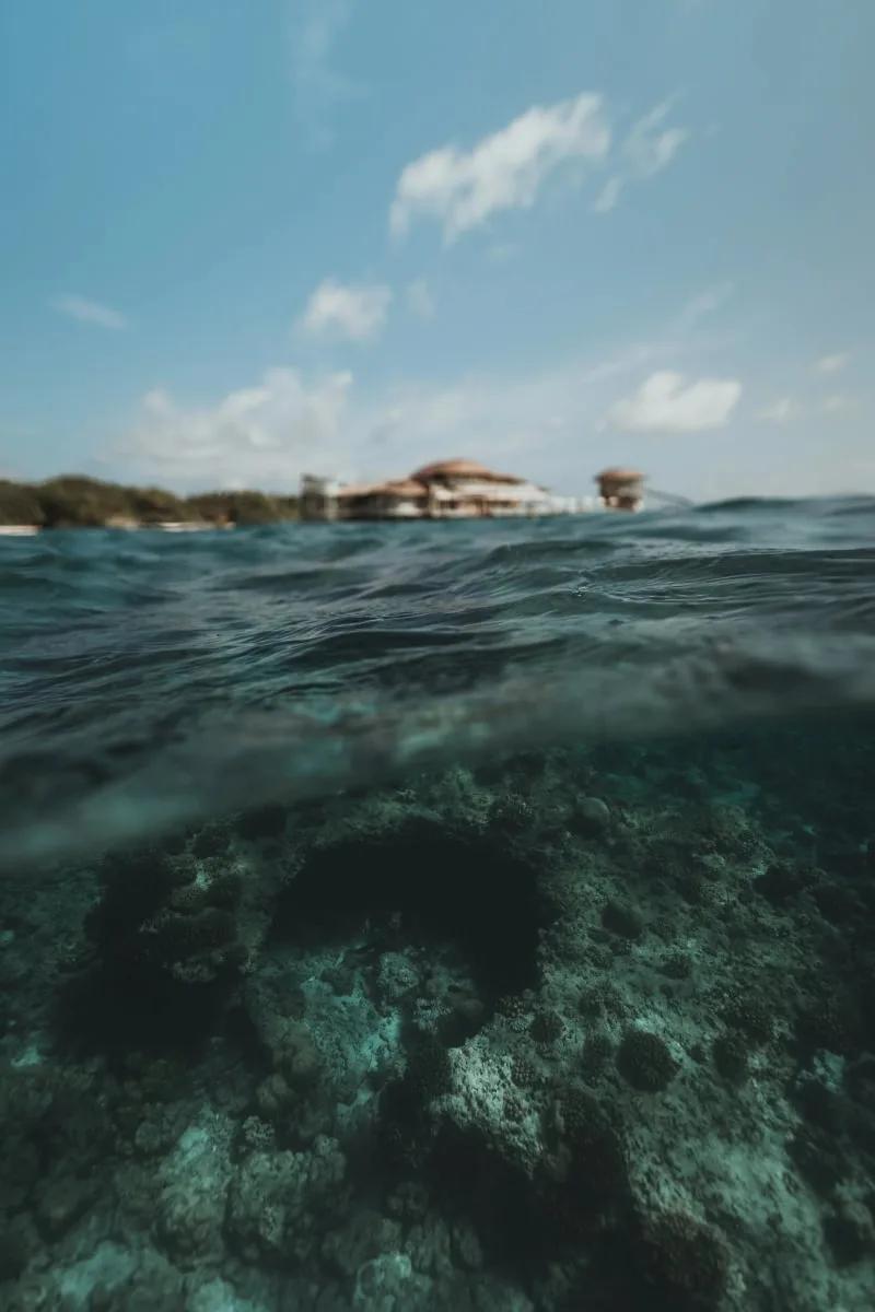 A split-level shot through the water's surface — coral structures visible underwater in the foreground, with Soneva Fushi's overwater villas on the horizon beyond