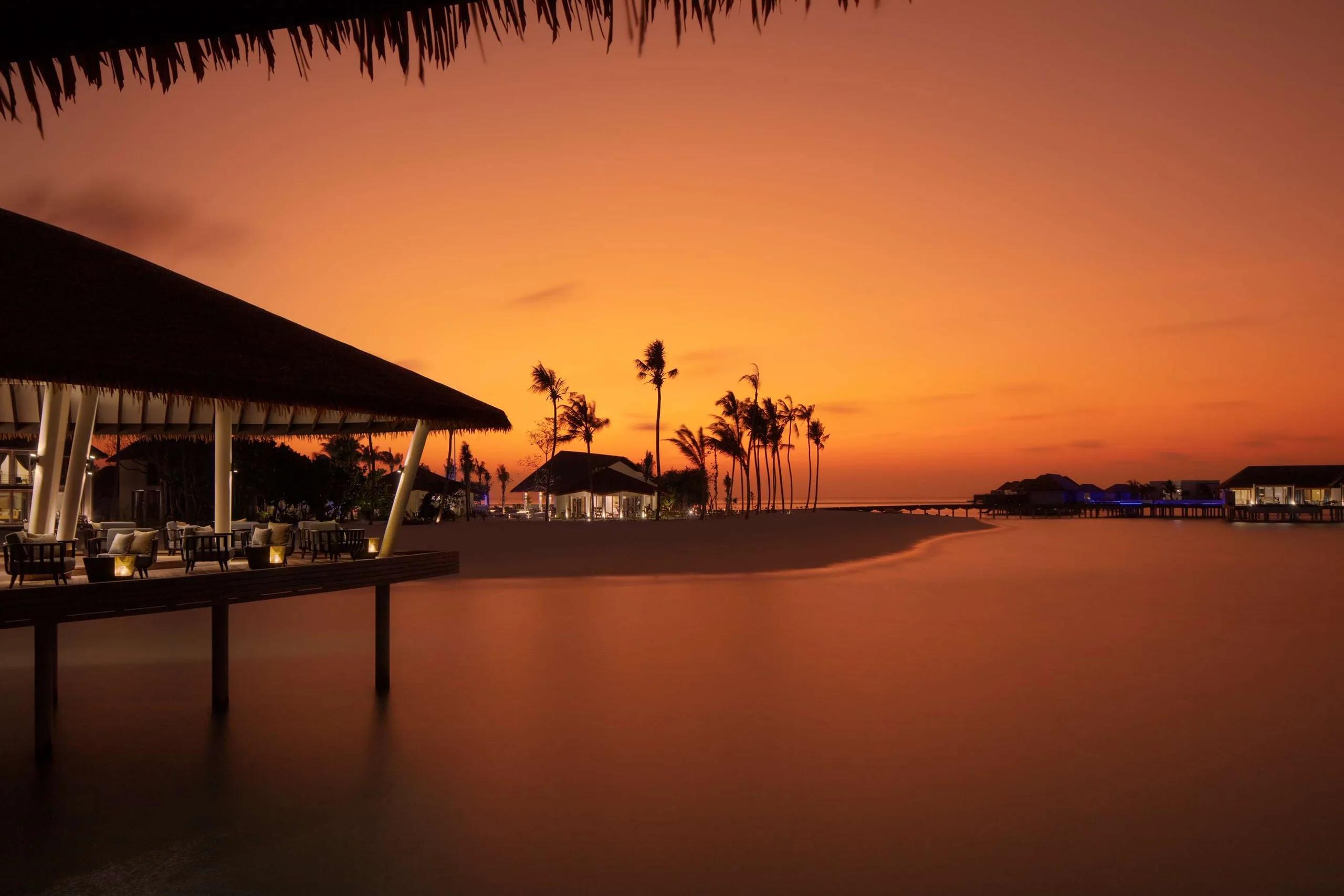 Mahurab Bar & Lounge's deck at dusk at Radisson Blu Resort Maldives, silhouetted palm trees and overwater villas along the darkening lagoon