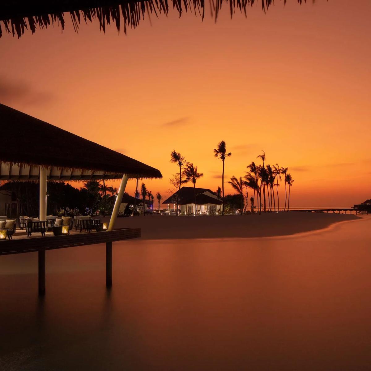 Mahurab Bar & Lounge's deck at dusk at Radisson Blu Resort Maldives, silhouetted palm trees and overwater villas along the darkening lagoon