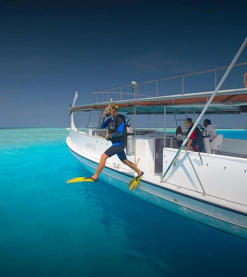 A scuba diver in fins stepping off the back of a Baros dive boat, the resort's name marked on the hull, into open water