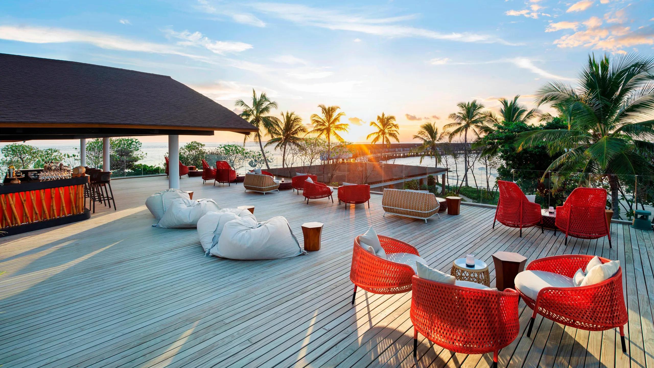 Sunset Bar's timber deck with red woven chairs, palm trees silhouetted against a setting sun