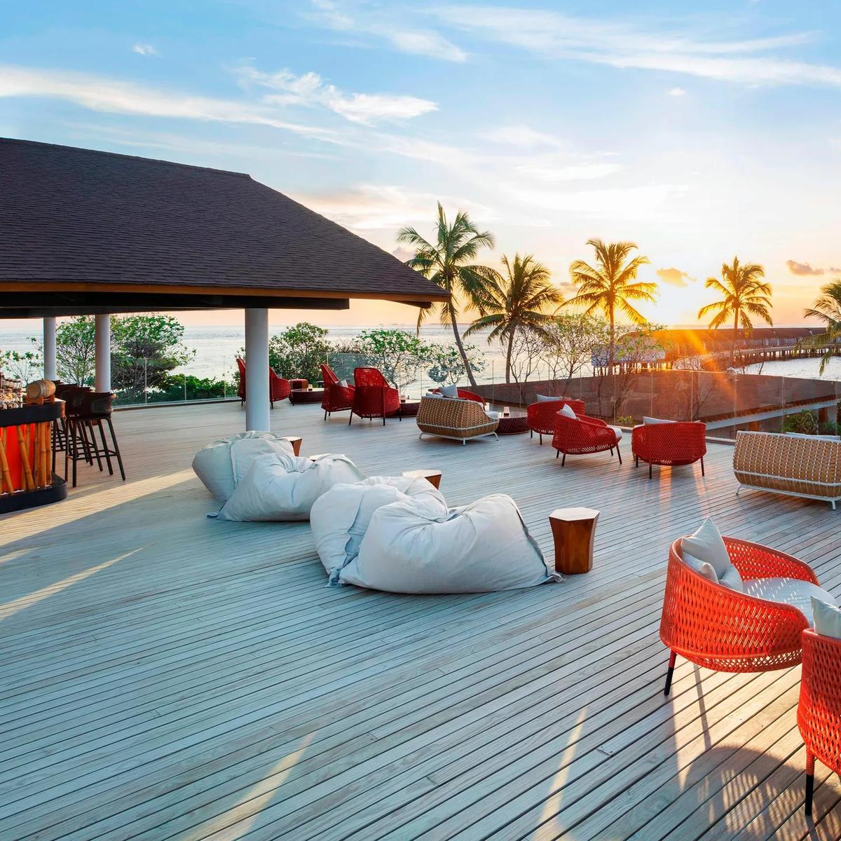 Sunset Bar's timber deck with red woven chairs, palm trees silhouetted against a setting sun