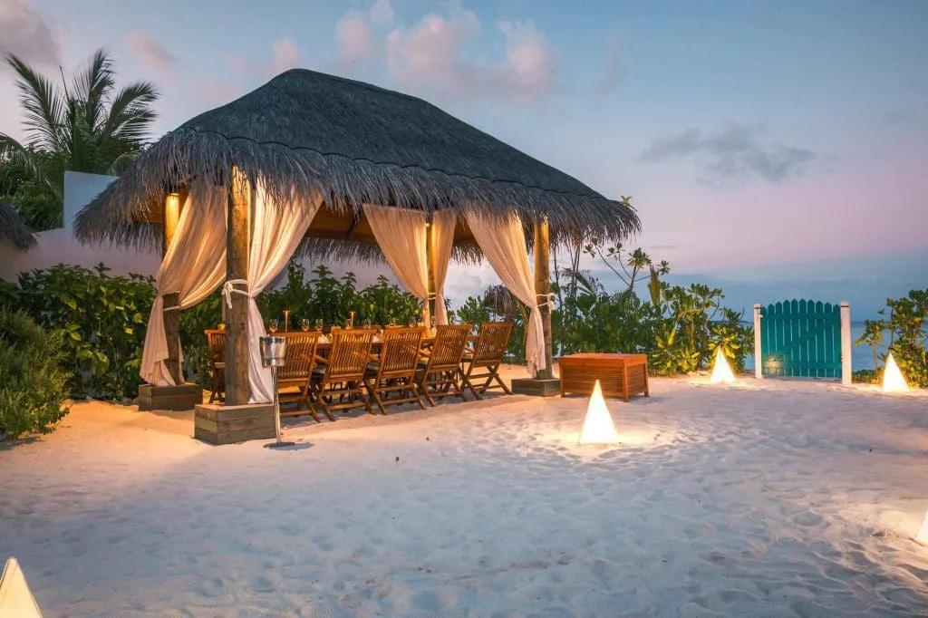 An open-air thatched cabana with tied-back curtains and a long dining table, set for the evening