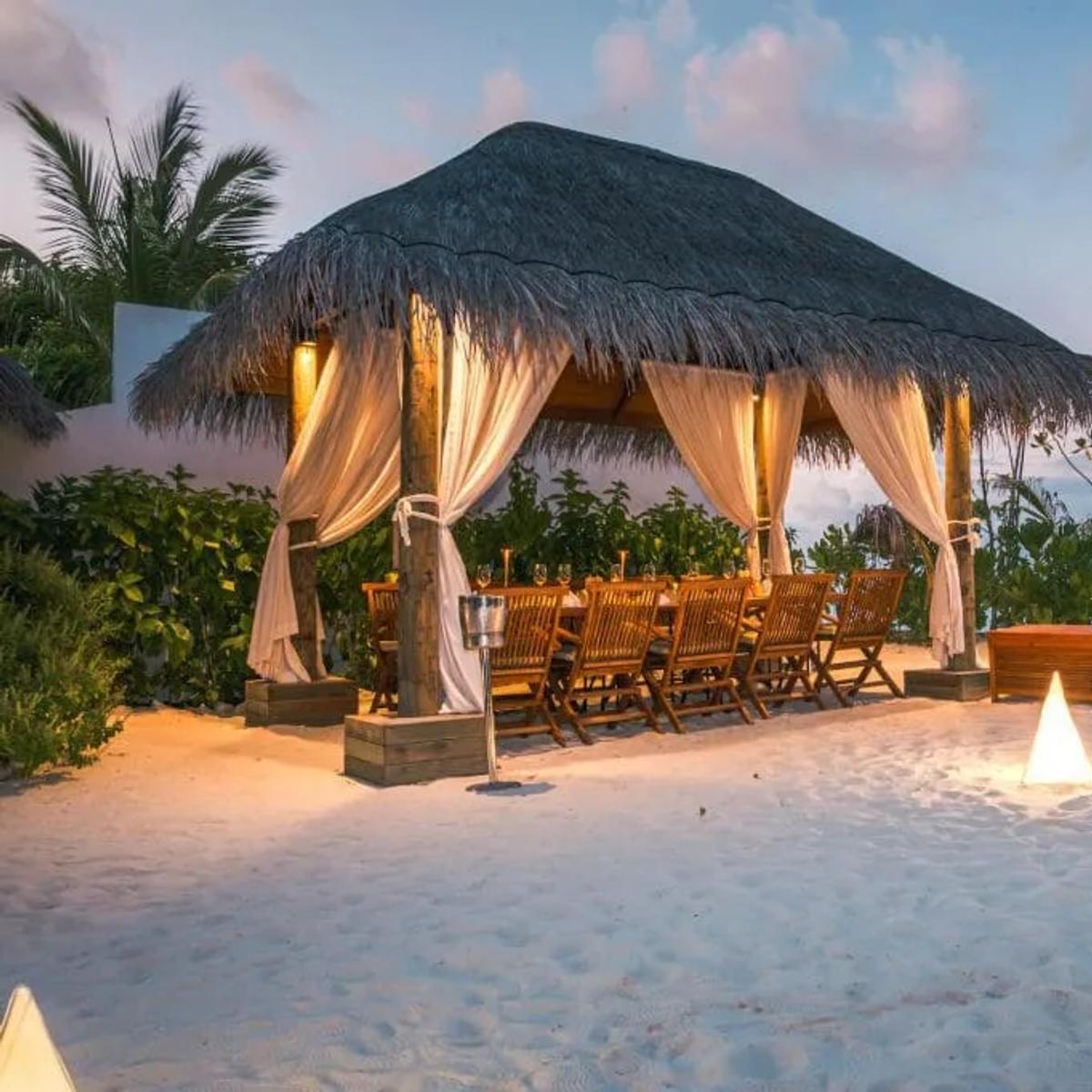 An open-air thatched cabana with tied-back curtains and a long dining table, set for the evening