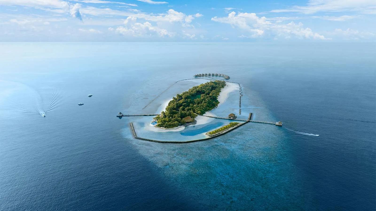 Aerial view of Komandoo's entire reef-ringed island, jetties reaching out to overwater villas on the northern tip and boats crossing the lagoon