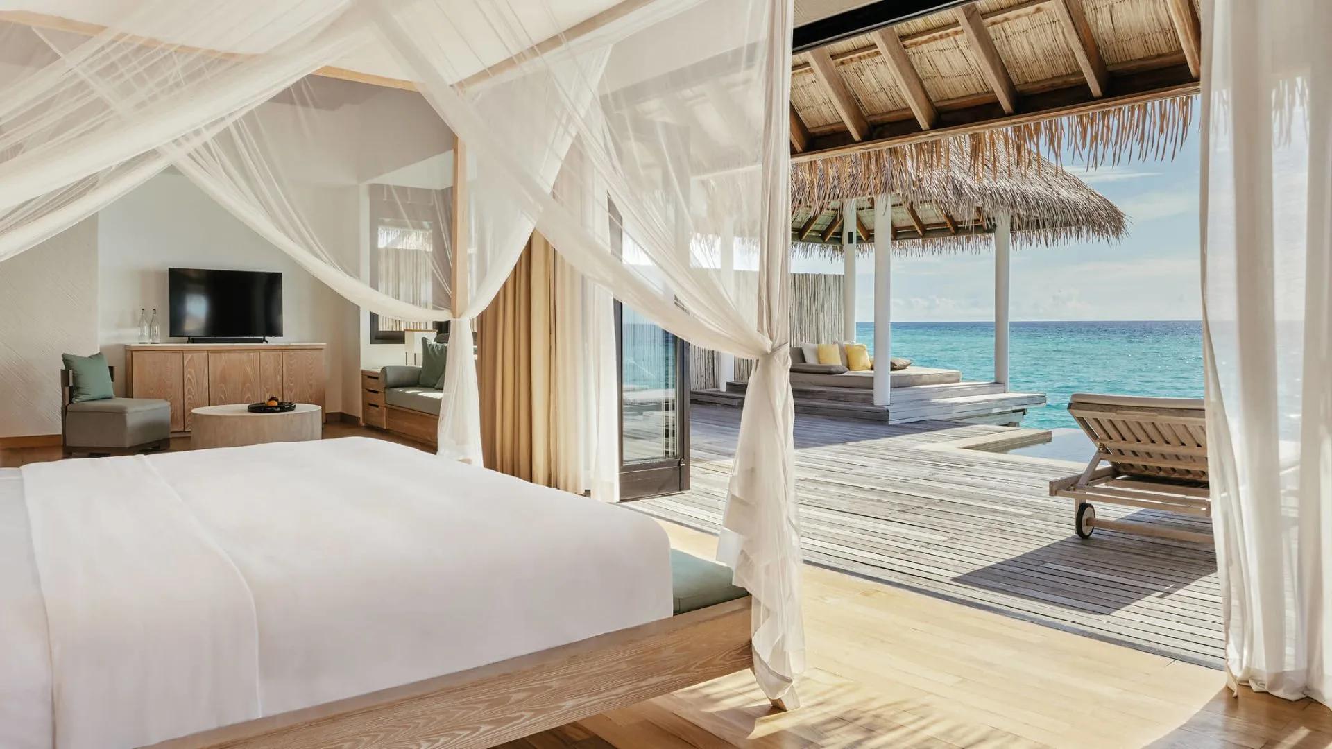 Water Suite bedroom with a canopy bed and TV console, doors open onto the overwater deck and daybed pavilion beyond