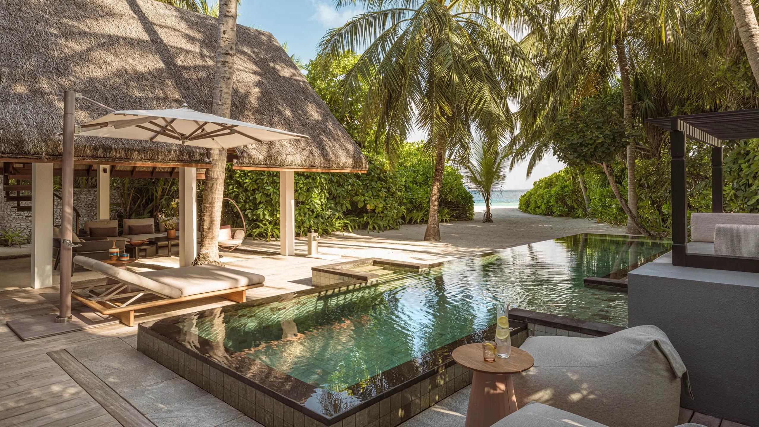 A Family Beach Villa's pool deck set under palm trees, with a thatched pavilion and the beach visible beyond