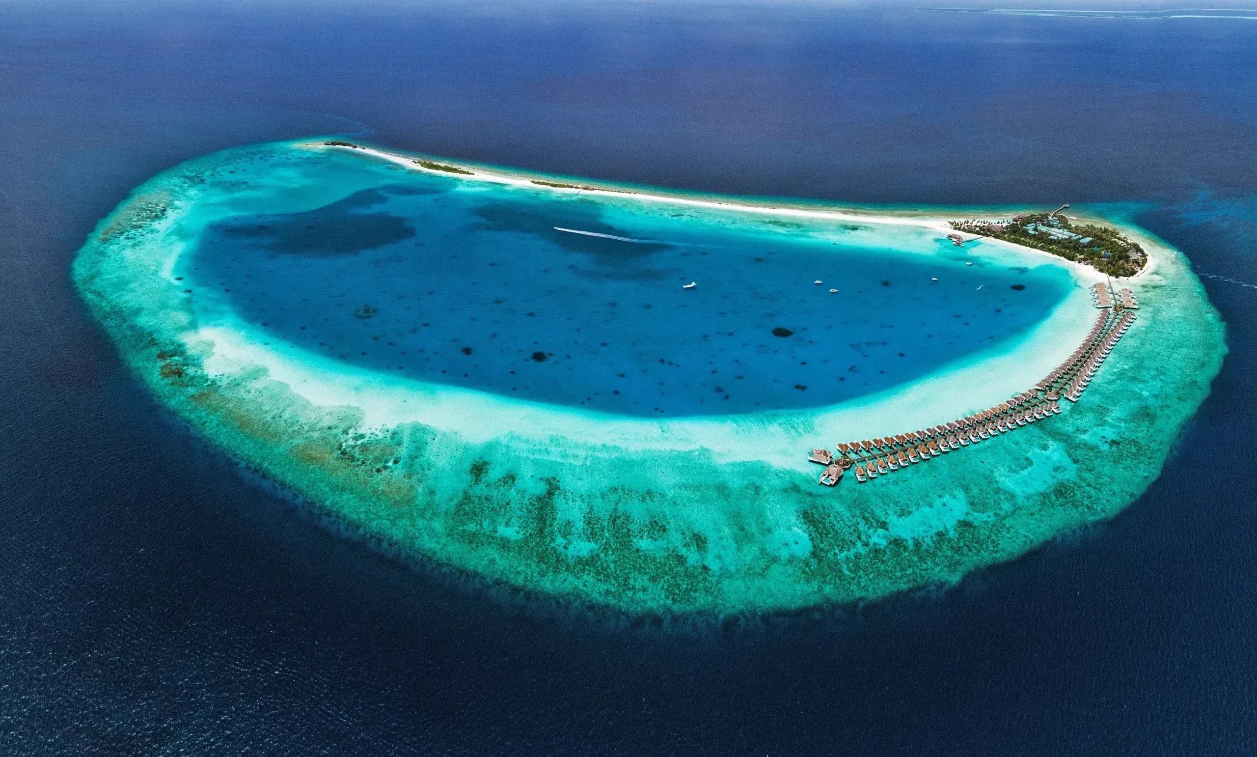 Aerial view of Finolhu's teardrop-shaped lagoon, ringed by reef, with white-sand beach along one edge and overwater villas on stilts extending from the island's tip