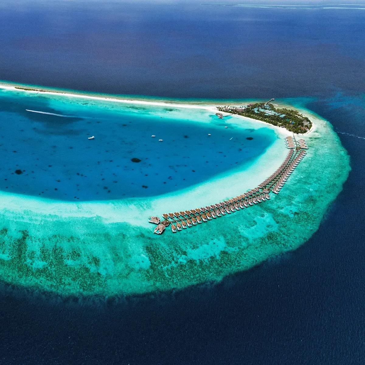 Aerial view of Finolhu's teardrop-shaped lagoon, ringed by reef, with white-sand beach along one edge and overwater villas on stilts extending from the island's tip