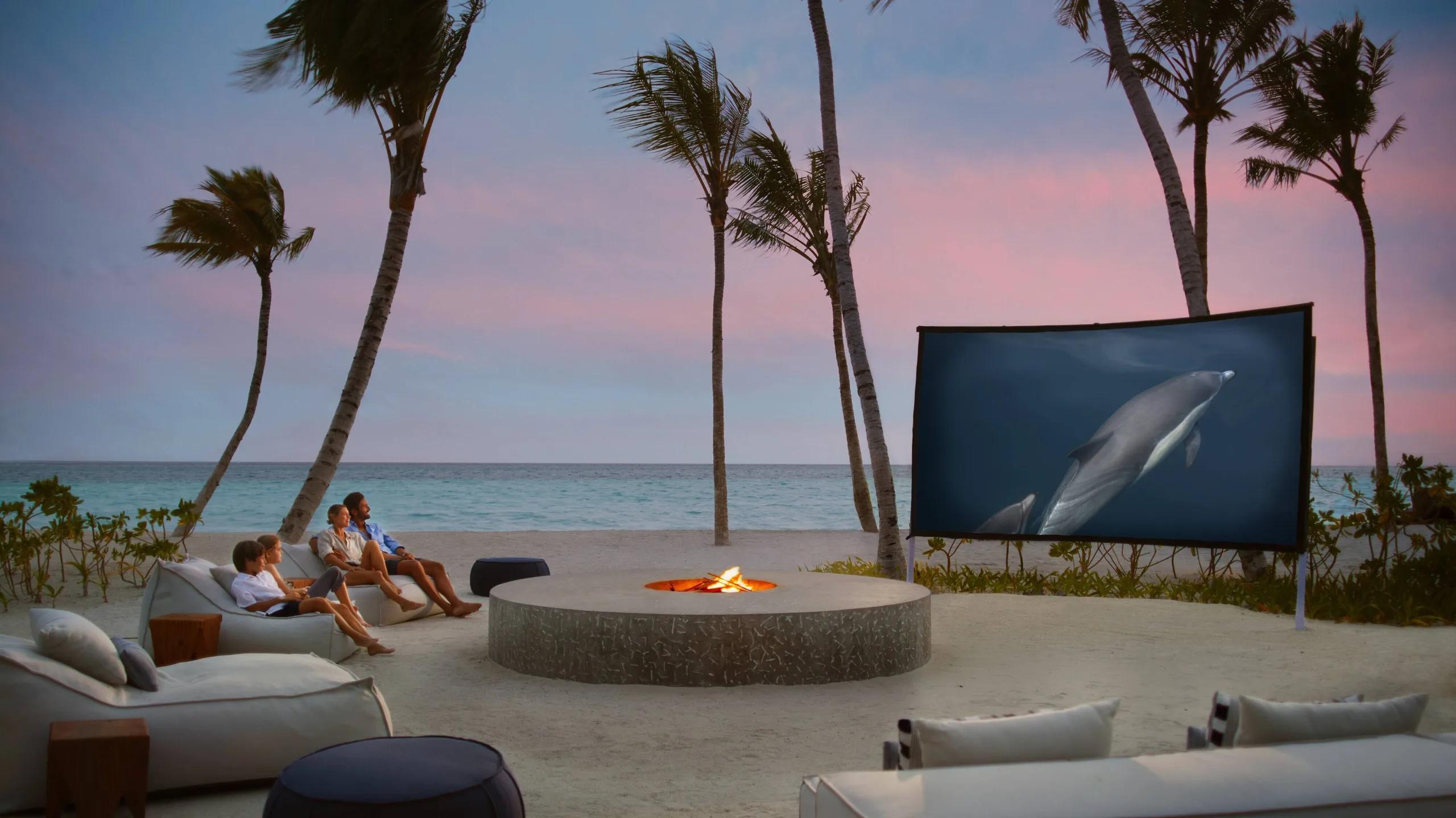 A family on beanbags beside a fire pit, watching an outdoor cinema screen on the beach at dusk