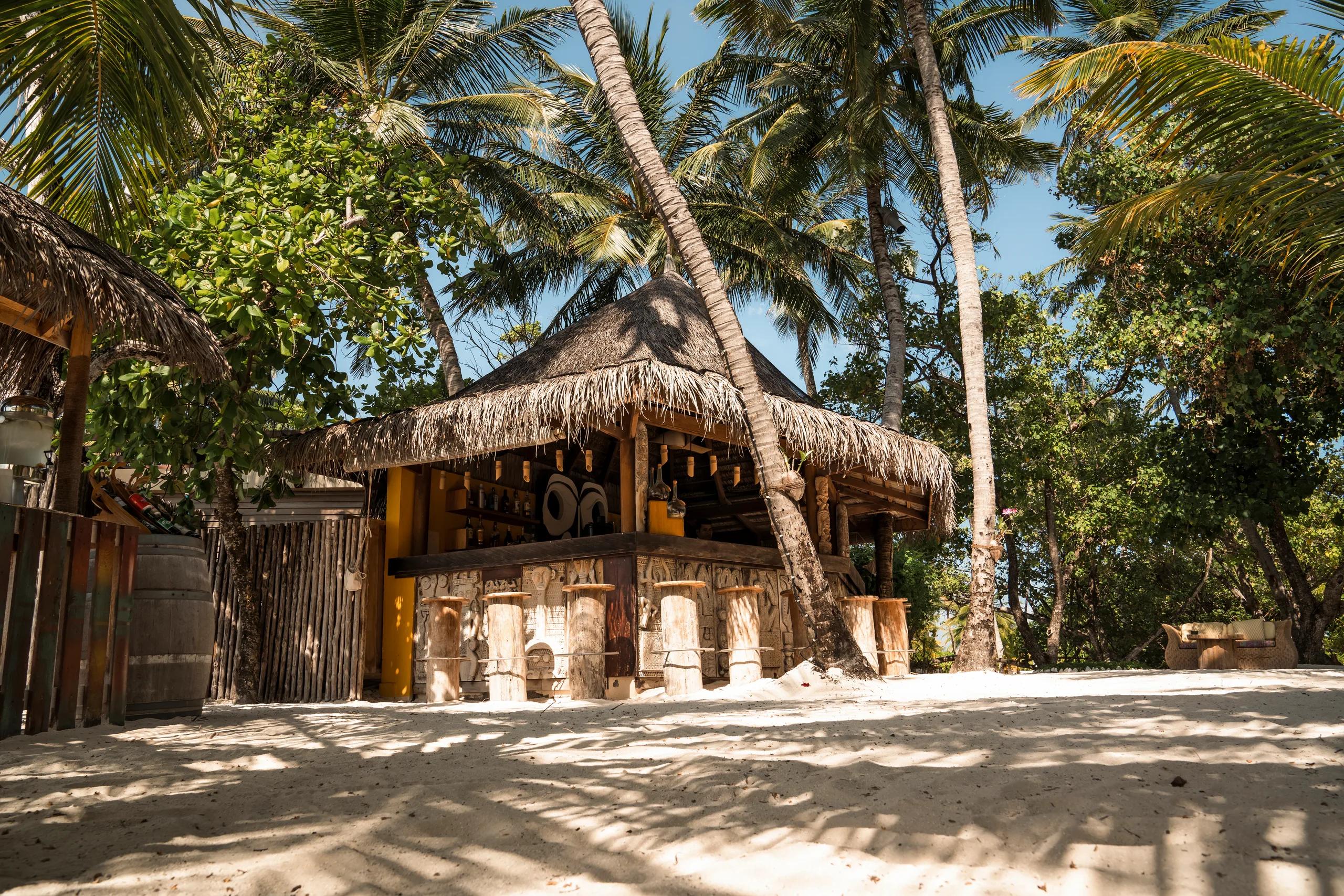 Totem Bar's driftwood-clad counter under a thatched roof, framed by leaning coconut palms