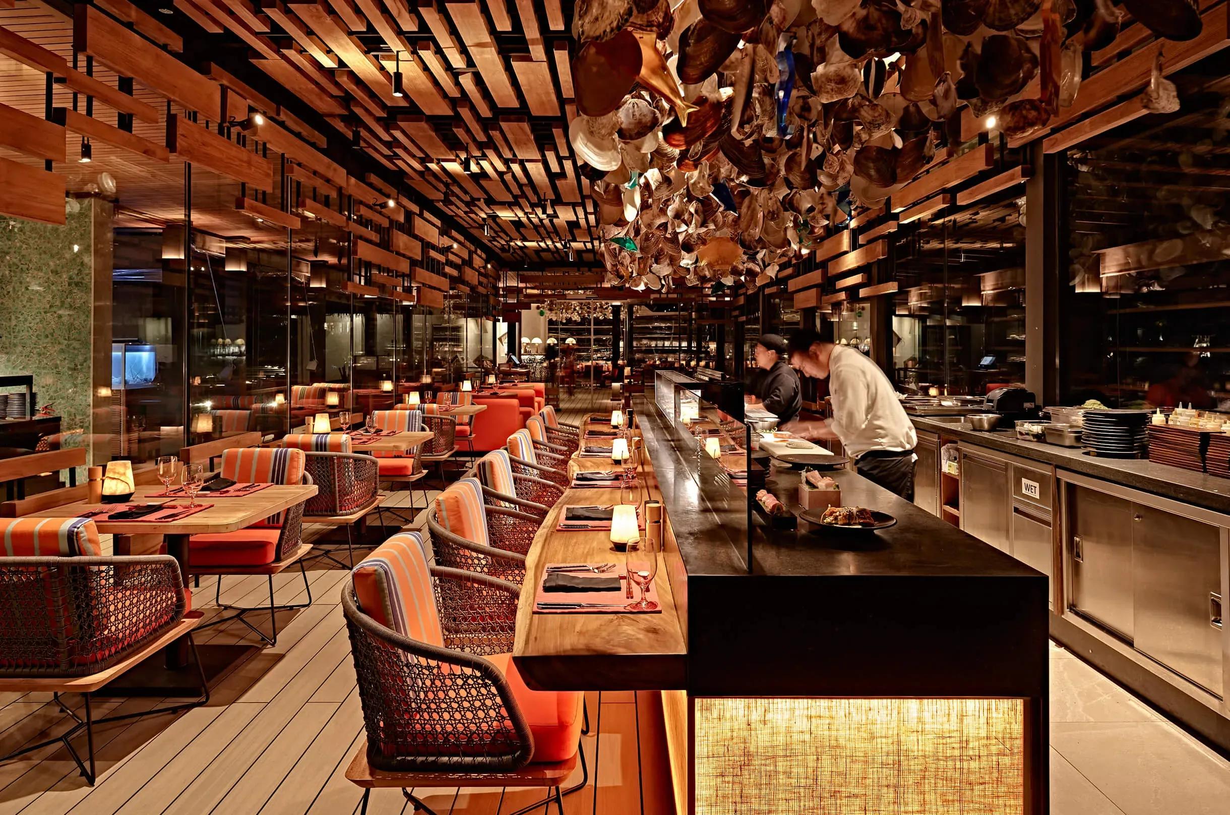 Kanusan's dining room, with a long sushi counter, orange-striped banquette seating and a ceiling installation of hanging oyster shells and glass above the chefs at work