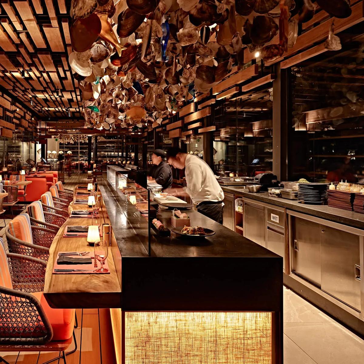 Kanusan's dining room, with a long sushi counter, orange-striped banquette seating and a ceiling installation of hanging oyster shells and glass above the chefs at work