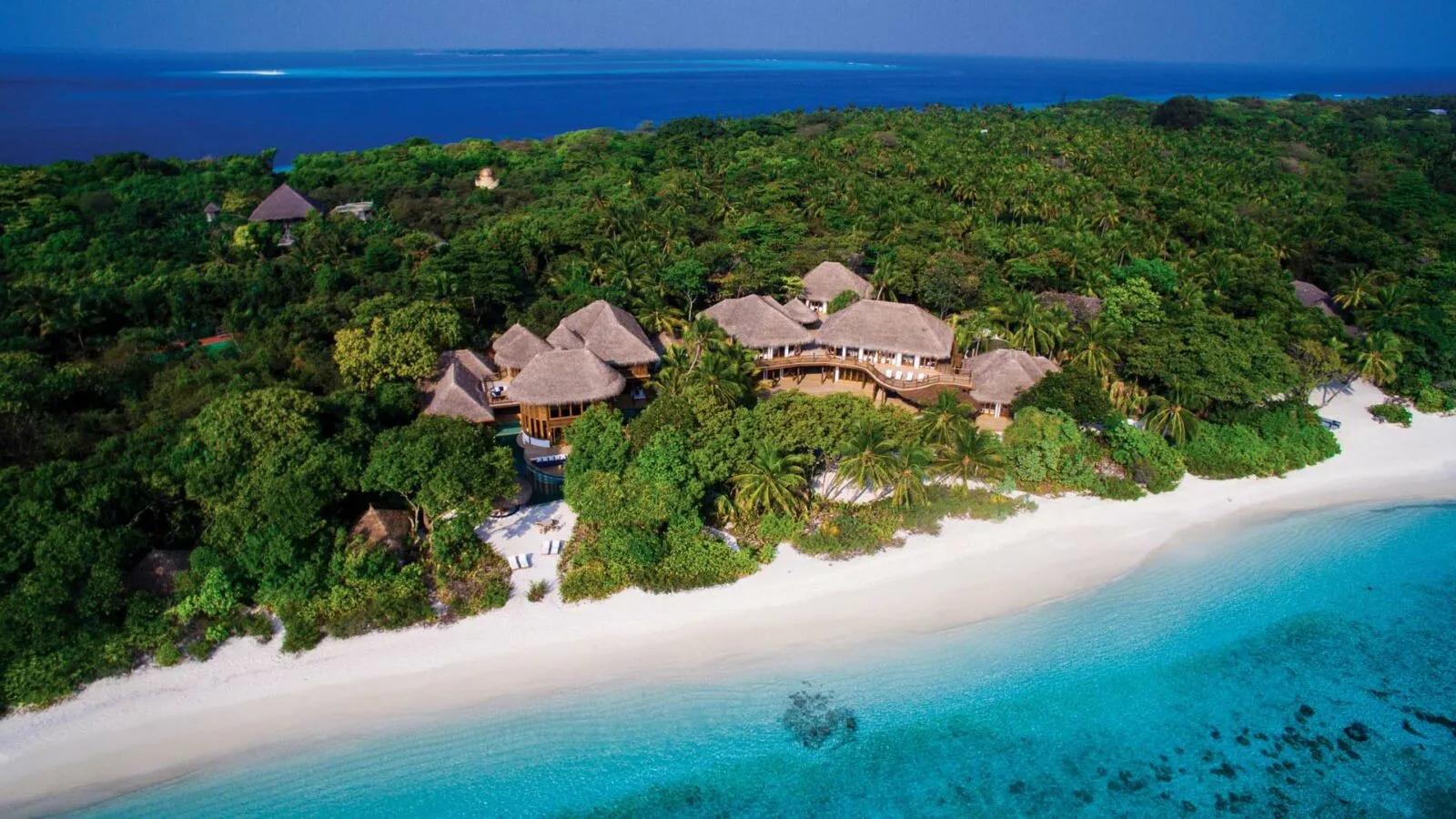 An aerial view of a beachfront villa complex at Soneva Fushi — thatched roofs and a private pool set among jungle, with a white-sand beach and the reef beyond