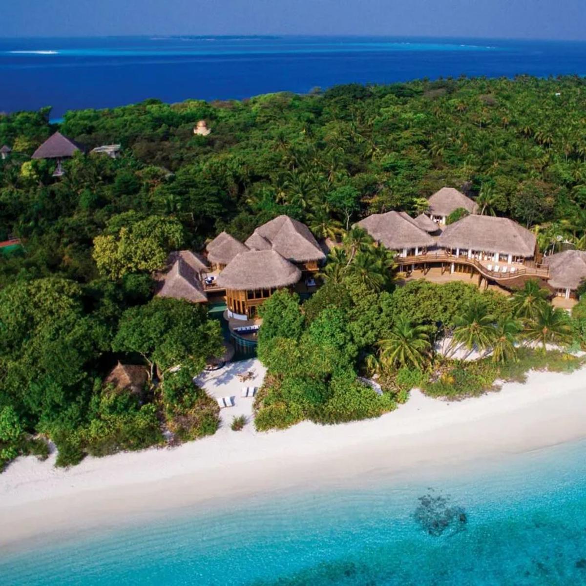 An aerial view of a beachfront villa complex at Soneva Fushi — thatched roofs and a private pool set among jungle, with a white-sand beach and the reef beyond