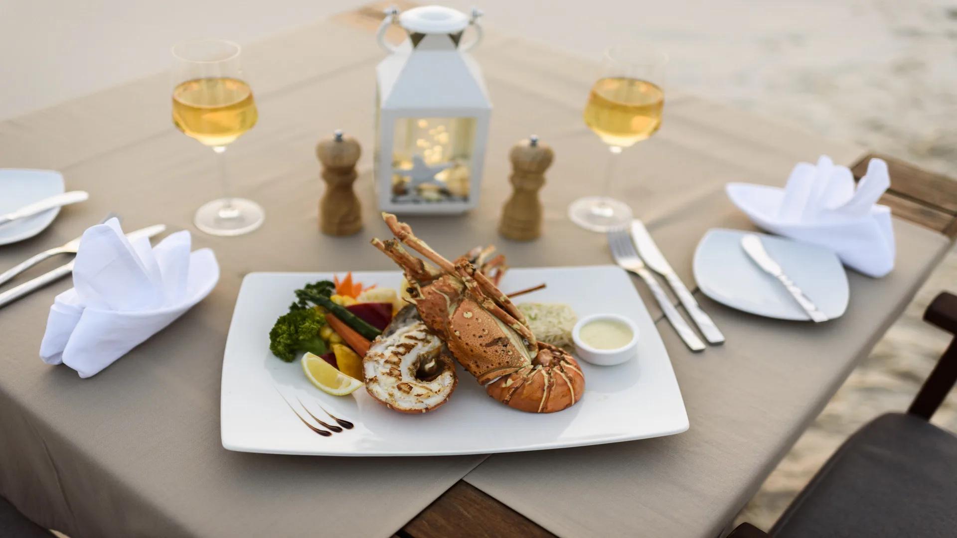 A grilled lobster dinner plated on a beachside table set with wine glasses and a lantern