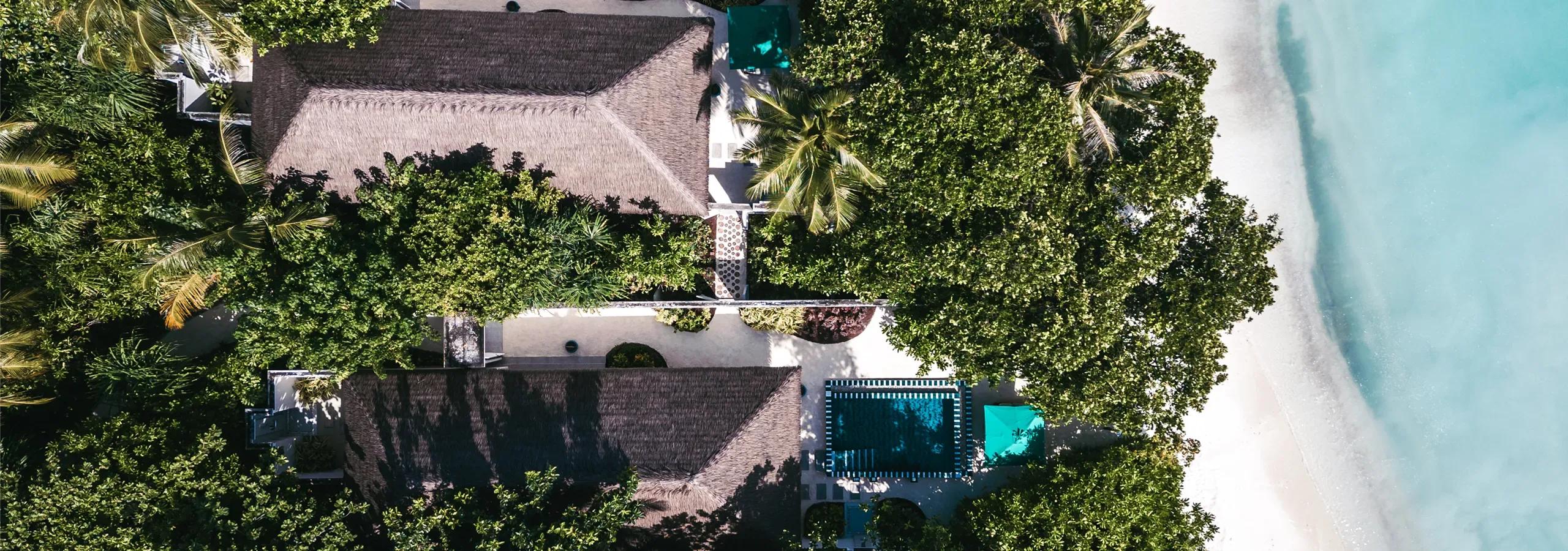 An aerial view of two thatched-roof Two Bedroom Interconnecting Beach Villas among the trees, each beside a tiled pool, a few steps from the beach